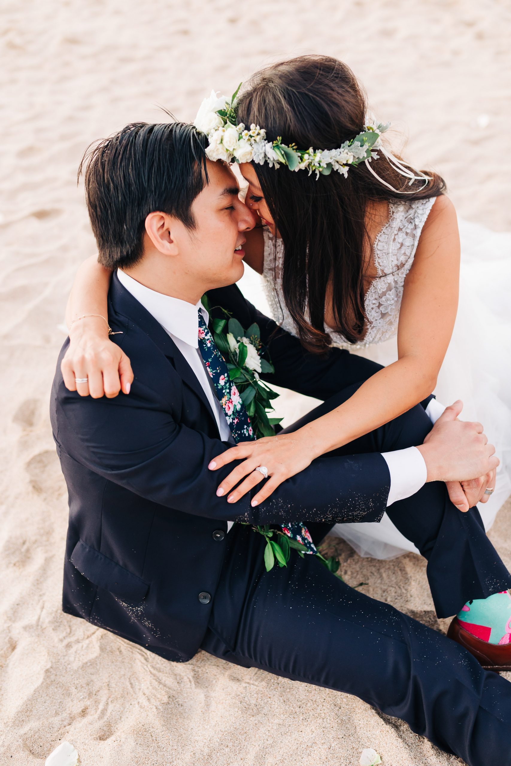 bride with flower crown hugging groom sitting in sand at beach wedding in Hawaii